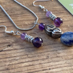 May include: A silver necklace and matching earrings set featuring amethyst and blue gemstone beads. The earrings have a hook closure. The jewellery is displayed on a wooden surface.