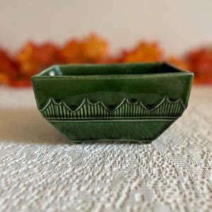 May include: A square, emerald green ceramic planter with a decorative, textured band around the middle. The planter sits on a white lace tablecloth, with blurred fall foliage in the background. The planter is approximately 4 inches wide.