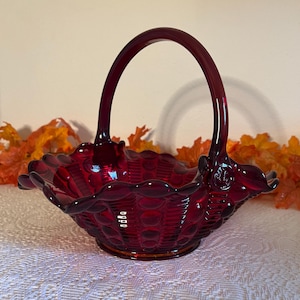 May include: A ruby red glass basket with a handle. The basket has a scalloped rim and a textured pattern. The handle is arched and attached to the sides of the basket. The basket is sitting on a white surface.