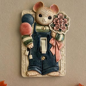 May include: A rectangular ceramic light switch plate featuring a sculpted mouse wearing blue overalls and a green striped shirt. The mouse holds two balloons and a bouquet of pink flowers. The switch toggle is in the center and labeled OFF.