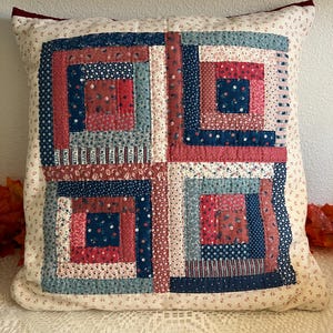 May include: A square throw pillow featuring a patchwork log cabin design. The center panel consists of four blocks made from various fabric strips in shades of red, navy blue, and light blue with floral and geometric patterns on a cream background.