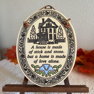May include: An oval ceramic wall plaque with a cream background and black border featuring floral motifs. It displays a black line drawing of a house and the text: A house is made of stick and stone, but a home is made of love alone. A blue flower sits below.