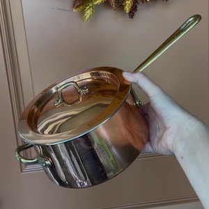 May include: A polished copper saucepan with a matching lid and brass handles. The pot features a rounded body, a long brass handle extending from the side, and a smaller brass handle on the lid. The surface is reflective and metallic in appearance.