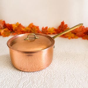 May include: A round copper saucepan with a matching lid and a long brass handle. The pot features a polished metallic finish and a small, curved brass handle attached to the center of the lid. The design is simple, functional, and circular.