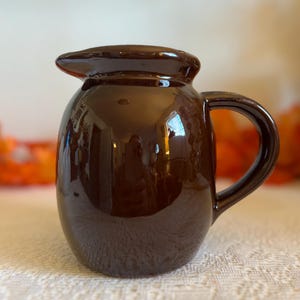 May include: A small, brown ceramic pitcher with a glossy finish. The pitcher has a rounded body, a curved handle, and a spout for pouring. The surface reflects light, and the pitcher sits on a white, textured surface. The overall design is simple and classic.