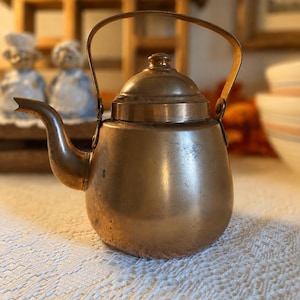 May include: A small, antique copper teapot with a curved spout and a handle. The teapot has a rounded body and a lid with a small knob. The copper has a warm, aged patina, suggesting it is vintage. The teapot is likely used for brewing tea.