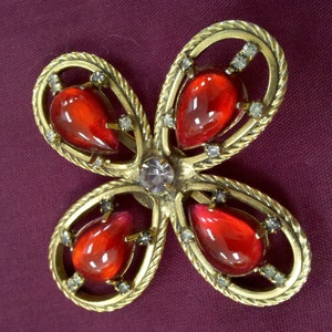 May include: A gold-tone brooch with a four-leaf clover design. Each leaf features a red teardrop-shaped stone and is surrounded by clear rhinestones.