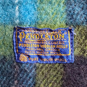May include: Close-up of a blue and green plaid fabric with a blue label. The label reads "Pendleton Woolen Mills, Portland, Oregon, 100% Virgin Wool". The fabric has a textured appearance.