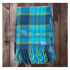 May include: A blue, green, and turquoise plaid blanket with fringe. The blanket has a label in the lower right corner. The blanket is folded and appears to be made of wool.