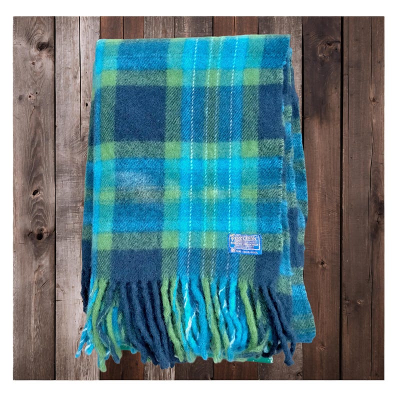 May include: A blue, green, and turquoise plaid blanket with fringe. The blanket has a label in the lower right corner. The blanket is folded and appears to be made of wool.