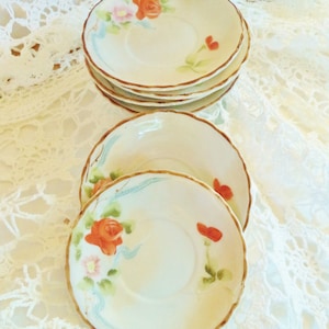 May include: A stack of vintage, hand-painted saucers. Each saucer features a cream-colored base with a scalloped edge and a brown trim. The saucers are decorated with orange roses, pink flowers, and blue accents.