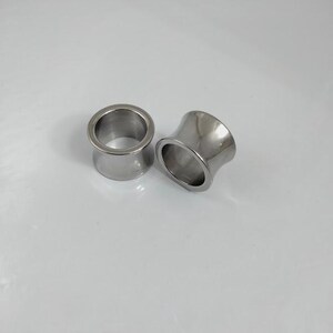 May include: Two polished, silver-colored ear gauges. The tunnels have a flared design, with a wider opening on each end. The gauges are made of a shiny, metallic material and are displayed on a white surface.