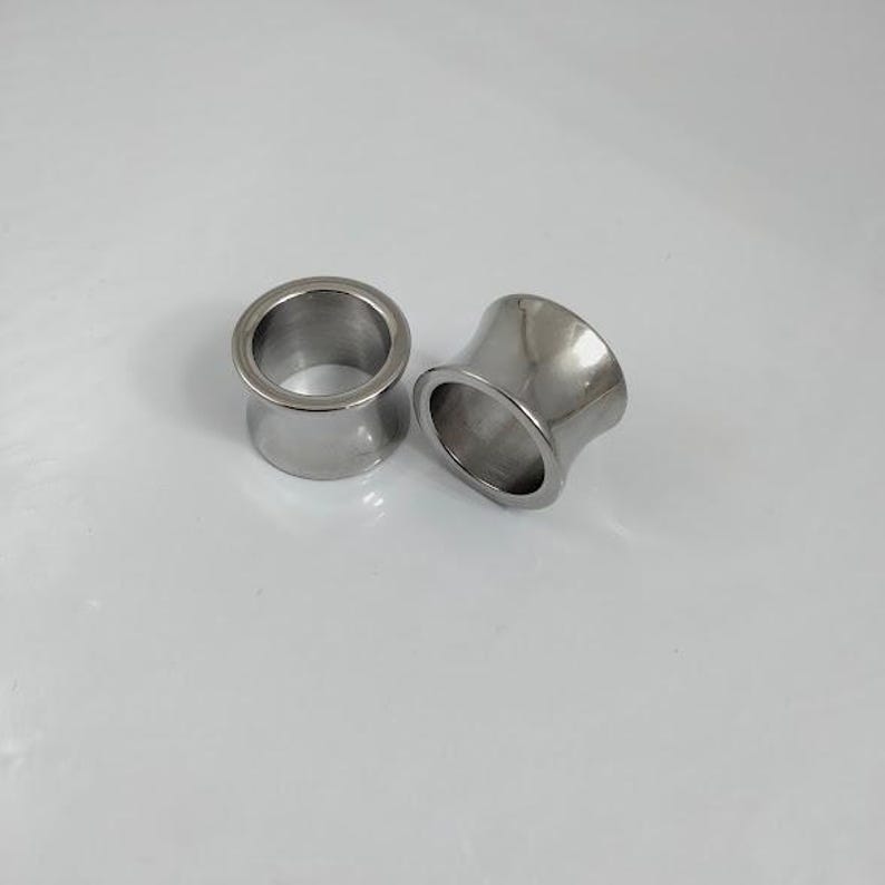 May include: Two polished, silver-colored ear gauges. The tunnels have a flared design, with a wider opening on each end. The gauges are made of a shiny, metallic material and are displayed on a white surface.