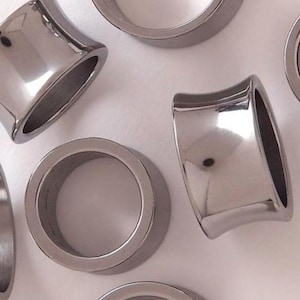 May include: Close-up of several silver-colored, flared ear gauges. The gauges have a polished, reflective surface and a rounded, cylindrical shape. The flared design widens towards the front and back, creating a unique look for body jewelry.