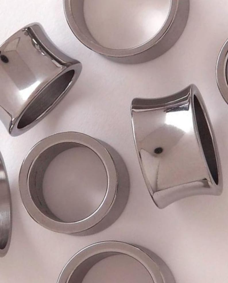 May include: Close-up of several silver-colored, flared ear gauges. The gauges have a polished, reflective surface and a rounded, cylindrical shape. The flared design widens towards the front and back, creating a unique look for body jewelry.