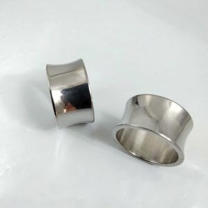 May include: Two polished, silver-colored ear gauges. The tunnels have a flared design, with a wider outer edge and a narrower center. The smooth, reflective surface of the jewelry catches the light, creating highlights and shadows.