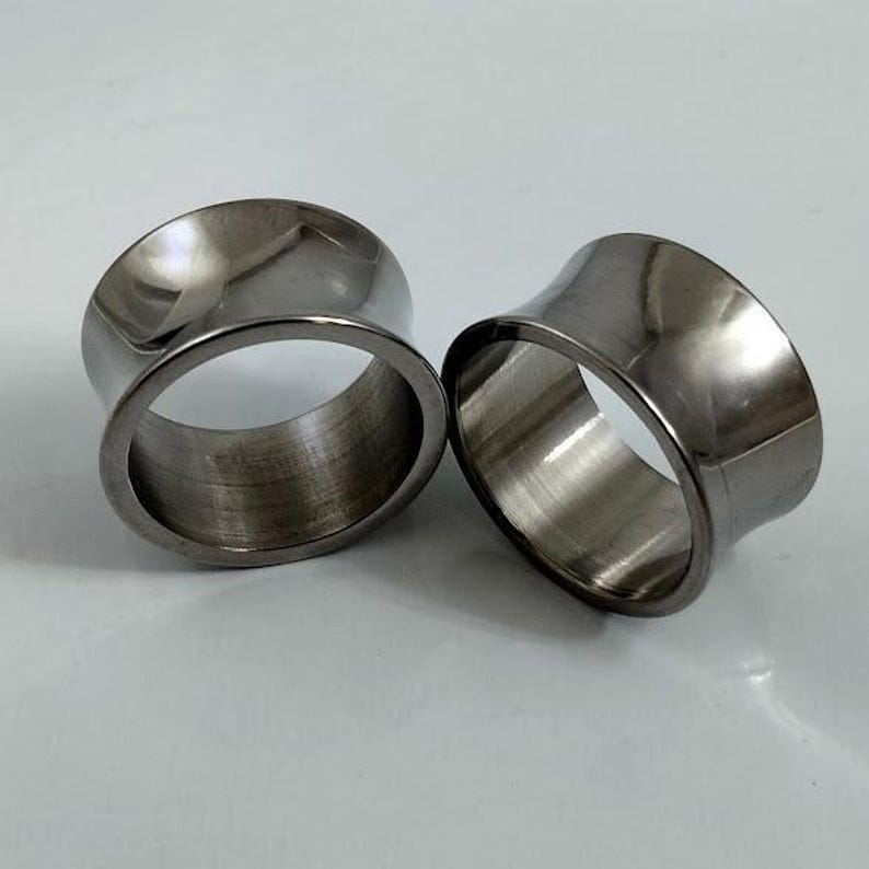 May include: Two polished, silver-toned ear gauges. The tunnels have a flared design, with a wider opening on one side. The gauges are made of a shiny, metallic material and are displayed on a white surface.