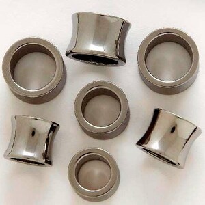 May include: Seven silver-toned, concave-shaped ear plugs are arranged on a white surface. The plugs have a polished, reflective finish, with a cylindrical center and flared edges. The design is symmetrical and modern, suitable for body modification.