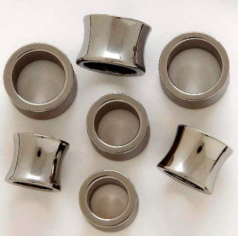 May include: Seven silver-toned, concave-shaped ear plugs are arranged on a white surface. The plugs have a polished, reflective finish, with a cylindrical center and flared edges. The design is symmetrical and modern, suitable for body modification.