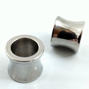 May include: Two polished, silver-colored ear gauges. The gauges have a flared design, with a wider outer rim and a narrower center. The smooth, metallic surface reflects light, creating highlights and shadows. The gauges are designed for stretched earlobes.