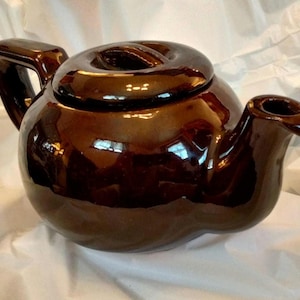 May include: A brown ceramic teapot with a rounded body and a lid with a unique design. The teapot has a spout and a handle.