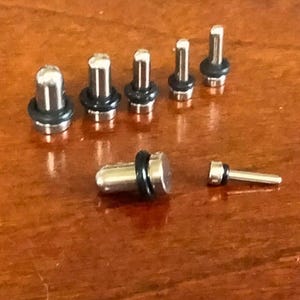 May include: A collection of silver-colored ear plugs with black rubber O-rings. The plugs vary in size, with a few laid out in a row and two separate. The plugs are on a brown wooden surface.