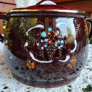 May include: A brown ceramic crock pot with a lid, decorated with gold and white floral designs and accents of orange and blue.