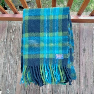 May include: A blue, green, and teal plaid scarf with fringe. The scarf has a rectangular shape and is made of a soft material. A small blue tag is sewn on the scarf. The scarf is folded and draped over a wooden railing.