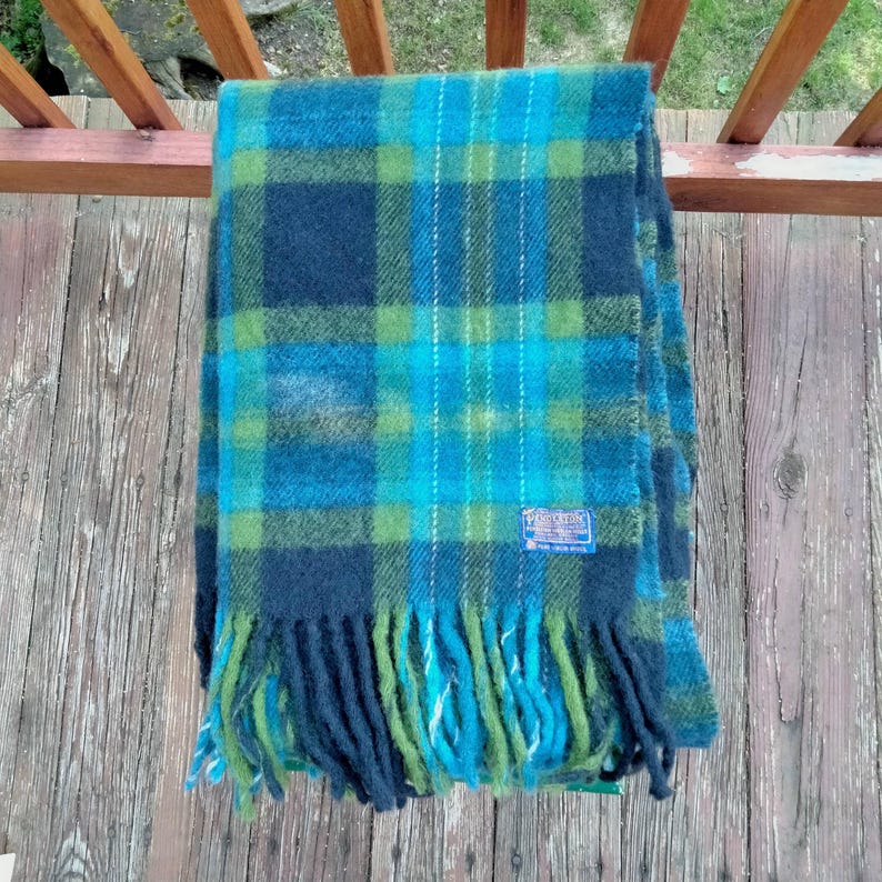 May include: A blue, green, and teal plaid scarf with fringe. The scarf has a rectangular shape and is made of a soft material. A small blue tag is sewn on the scarf. The scarf is folded and draped over a wooden railing.
