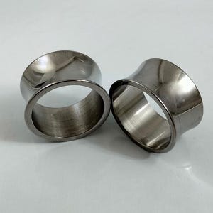 May include: Two polished, silver-toned ear gauges. The tunnels have a flared design, with a wider outer rim and a narrower inner rim. The gauges are made of a shiny, metallic material and are displayed on a light background.