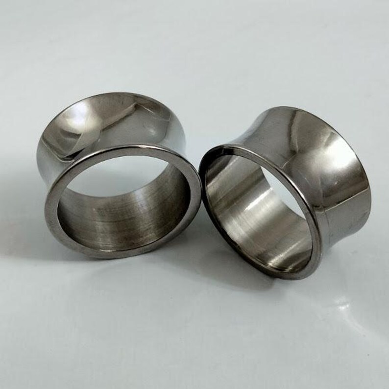 May include: Two polished, silver-toned ear gauges. The tunnels have a flared design, with a wider outer rim and a narrower inner rim. The gauges are made of a shiny, metallic material and are displayed on a light background.