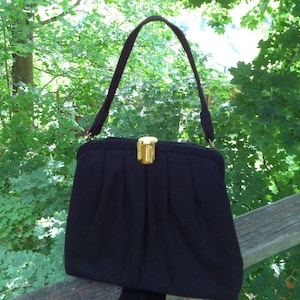 May include: A black handbag with a gold clasp and a handle. The purse has a pleated design and a rectangular shape. The background is a blurred view of green foliage and a wooden fence.