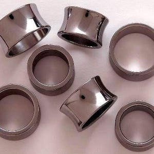 May include: A collection of dark gray, metallic ear gauges in various shapes. Some are flared, while others are cylindrical. The gauges are arranged on a light surface, reflecting the surrounding light.