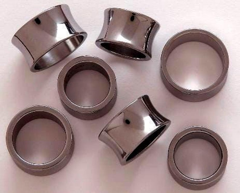 May include: A collection of dark gray, metallic ear gauges in various shapes. Some are flared, while others are cylindrical. The gauges are arranged on a light surface, reflecting the surrounding light.