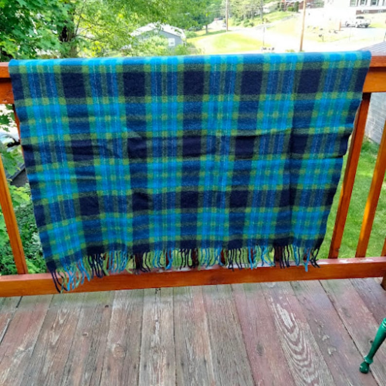 May include: A blue, green, and black plaid blanket with fringe. The blanket is draped over a wooden railing on a deck. The plaid pattern is a classic design, and the fringe adds a decorative touch.
