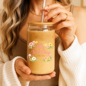 Puede incluir: Un vaso de vidrio transparente con tapa de bambú y pajita transparente, lleno de una bebida marrón claro. El vaso presenta un diseño floral y el texto "This Is the Day" con el versículo bíblico Salmo 118:24.