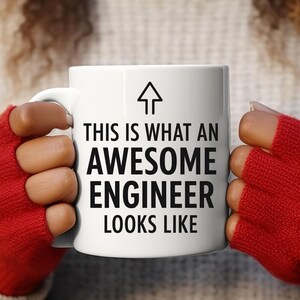 以下が含まれることがあります： 「THIS IS WHAT AN AWESOME ENGINEER LOOKS LIKE」の黒い文字と矢印のグラフィックが入った白いセラミックマグ。赤い指なし手袋をはめた人がマグカップを持っています。