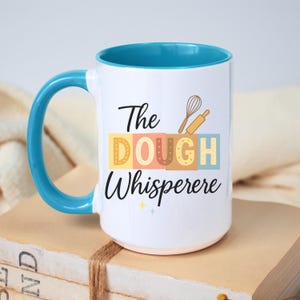 May include: White ceramic mug with a teal interior and handle. The mug features the text "The DOUGH Whisperere" in a whimsical font, with colorful block letters and baking-themed illustrations. The mug is sitting on a stack of books.