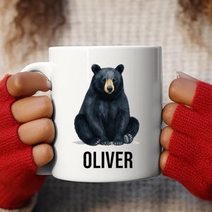 May include: White ceramic mug with a black bear illustration and a tan snout. The name "OLIVER" is printed in black block letters below the bear. The mug is held by a person wearing red fingerless gloves.