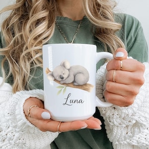 May include: White ceramic mug featuring a sleeping koala bear illustration on a branch with green leaves. The mug has the name "Luna" printed in a script font. The mug has a large handle and is held by a person.