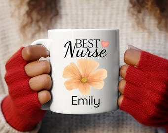 May include: White ceramic mug with the words "BEST Nurse" in black script, a peach-colored flower, and the name "Emily". The mug is held by a person wearing red gloves.