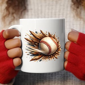 May include: White ceramic mug featuring a baseball design. The baseball is depicted with red stitching and a brown and gold burst effect. The mug is held by a person wearing red gloves.