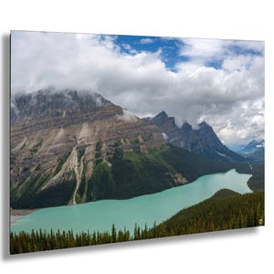Peut inclure: Impression d'un paysage pittoresque avec un lac turquoise bordé de montagnes et de forêts de conifères. Le ciel est rempli de nuages blancs, créant un contraste saisissant avec le terrain accidenté. L'impression est encadrée en noir.