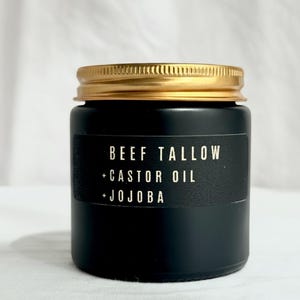 May include: A black jar with a gold lid. A black label on the jar reads "BEEF TALLOW + CASTOR OIL + JOJOBA" in white text. The jar is sitting on a white surface.