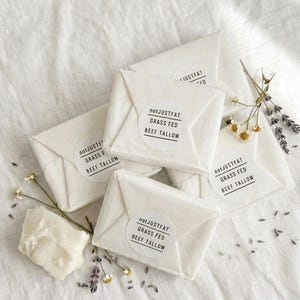 Puede incluir: Varios paquetes envueltos en papel blanco con etiquetas circulares que dicen "notJUSTFAT GRASS FED BEEF TALLOW". También hay un trozo de sebo y ramitas de flores. Los artículos están dispuestos sobre una tela blanca.