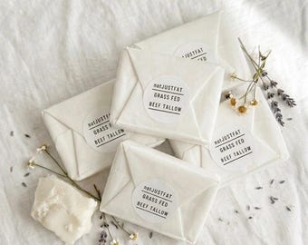 May include: Several white paper-wrapped packages with circular labels that read "notJUSTFAT GRASS FED BEEF TALLOW." A chunk of tallow and sprigs of flowers are also present. The items are arranged on a white cloth.