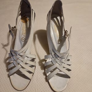 May include: A pair of white sandals featuring a woven strap design across the front, an ankle strap with a metal buckle, and a heel. The interior sole is marked with the text #546, the brand name, and the size 39.