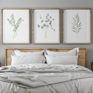 Può includere: Una scena di camera da letto con un letto con biancheria da letto bianca e una coperta grigia. Sopra il letto ci sono tre stampe botaniche incorniciate con illustrazioni ad acquerello verde. Comodini in legno e lampade a sospensione completano l'arredamento.