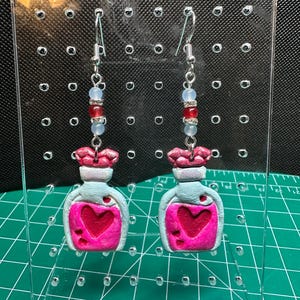 May include: A pair of handmade earrings featuring small, light blue potion bottle charms with pink heart designs. The charms are topped with pink lips and accented with red and blue beads on silver-tone hooks.