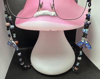 May include: Eyeglasses with a navy blue frame and pink lenses rest on a pink mushroom-shaped stand. A beaded eyeglass chain with blue and black beads and blue moth charms hangs from the glasses. The background is black.